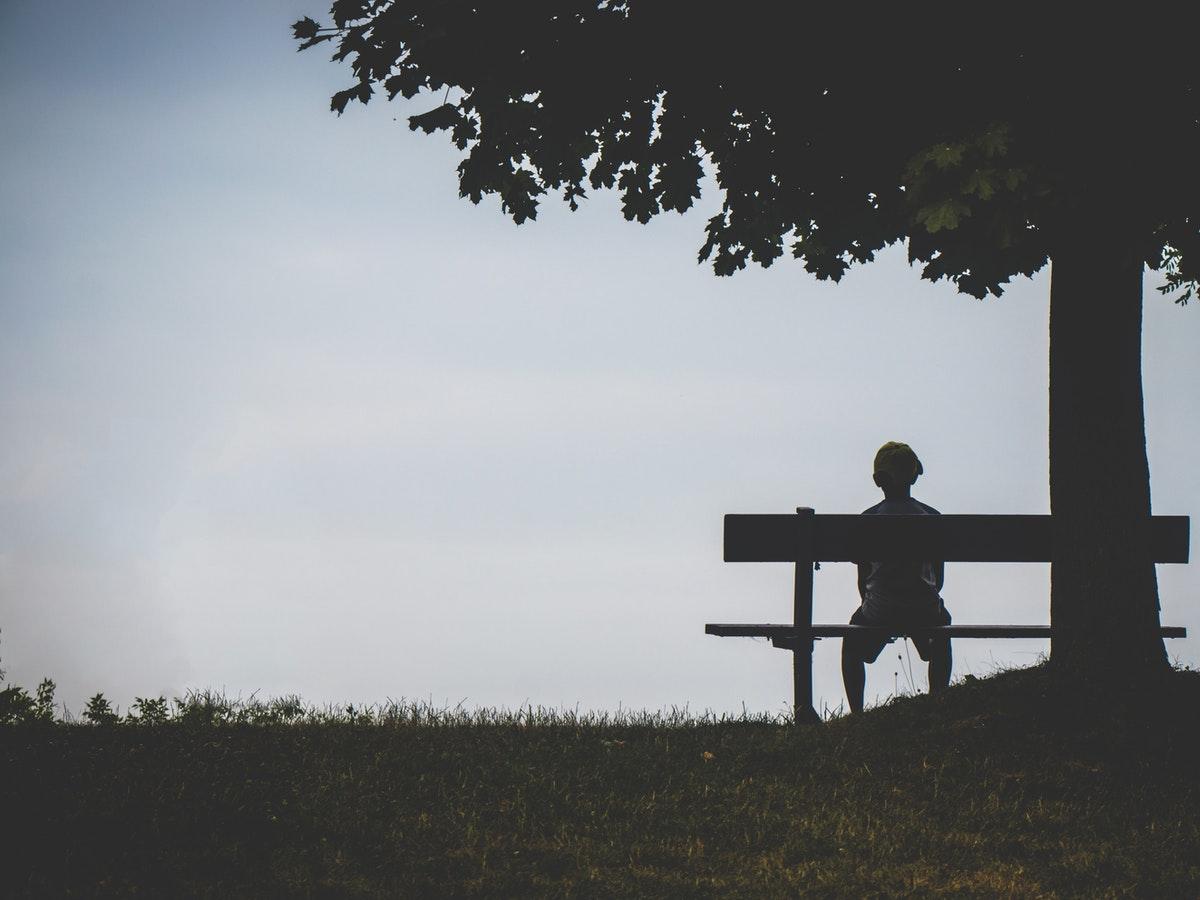 A lonely person sitting under a tree on a bench and looking in the distance