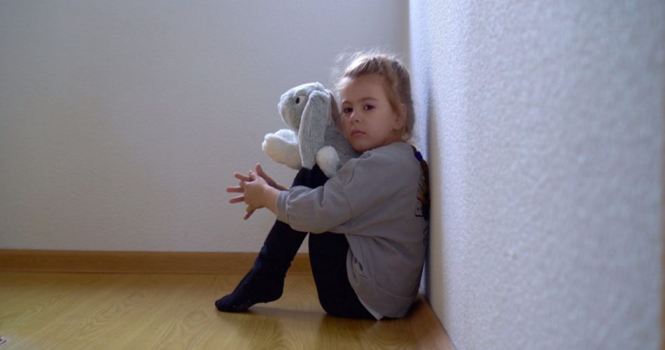 A sad looking child sitting in the corner with a stuff toy in her hand