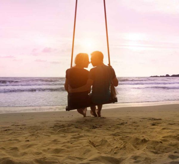 A couple in relationship sitting together on a swing on the beach during the sunset